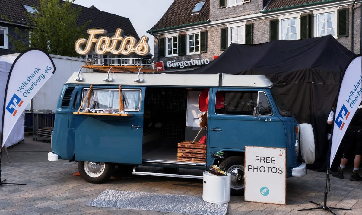 Der blaue Foto Bulli Ulli auf einem Ortsfest, links und rechts je ein Beachflag der Volksbank Oberberg eG, davor ein Schild mit der Aufschrift FREE PHOTOS