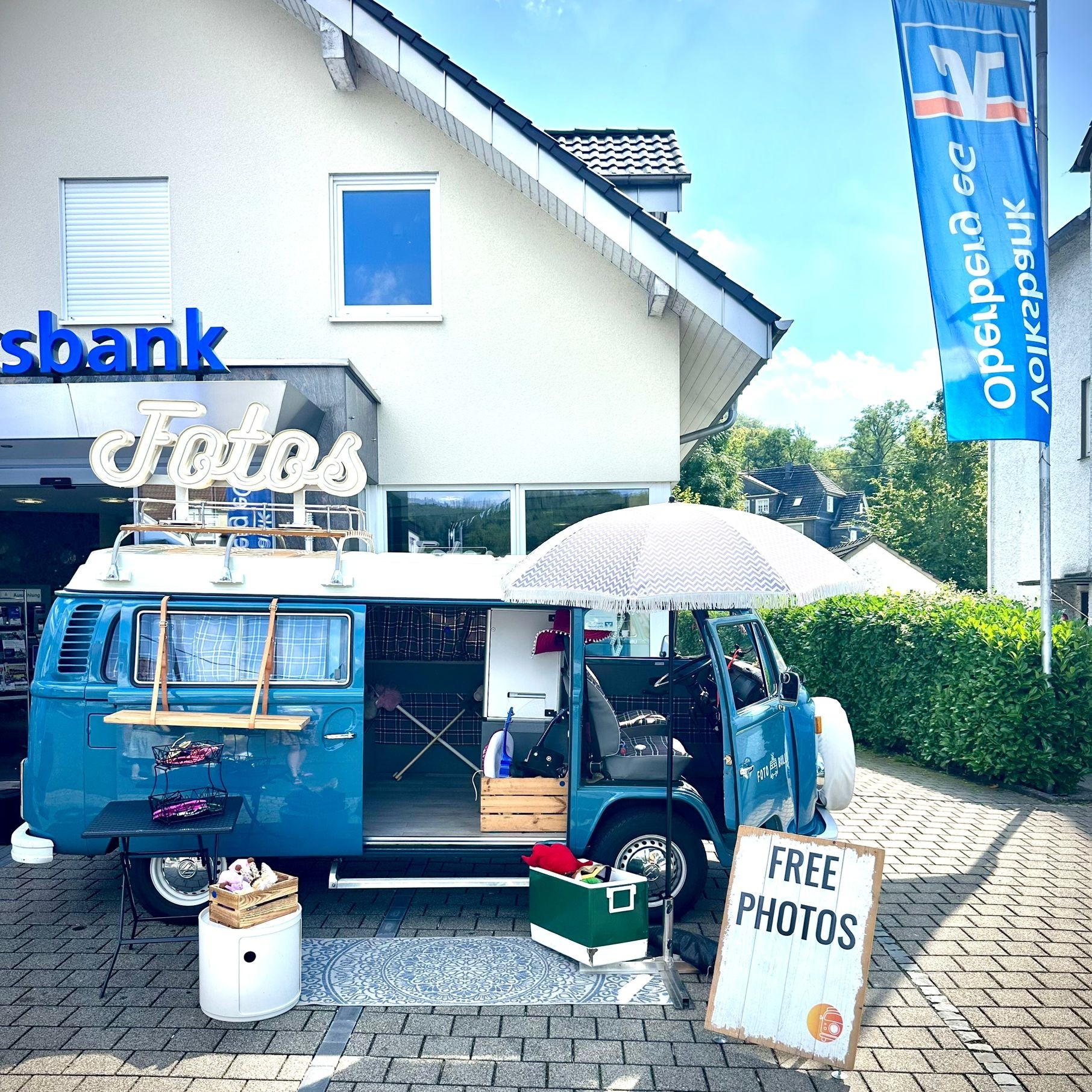 Der blaue Foto Bulli Ulli steht an einem Aktionstag direkt vor einer Filiale der Volksbank Oberberg eG, daneben ein Schild mit der Aufschrift FREE PHOTOS