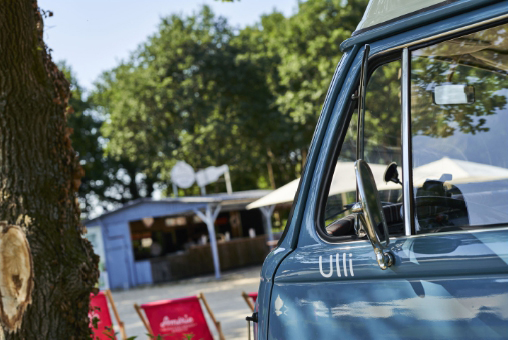 Nahaufnahme der Fahrerseite des blauen Foto Bullis mit dem Schriftzug Ulli, im Hintergrund Liegestühle und eine Strandbar